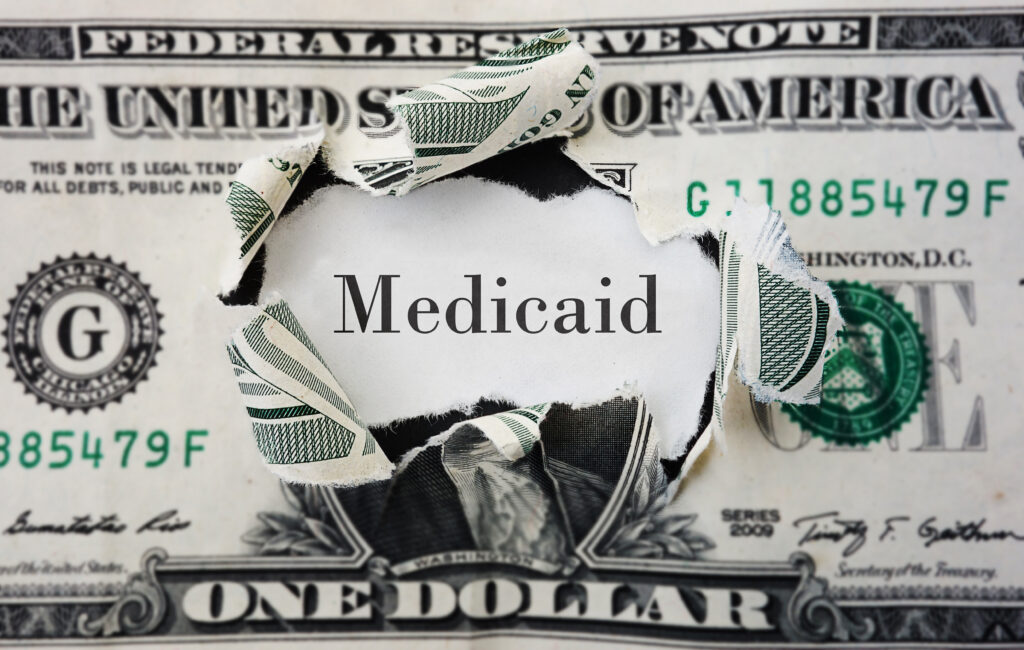 Hole in dollar bill revealing the word "Medicaid." Healthcare fraud concept.