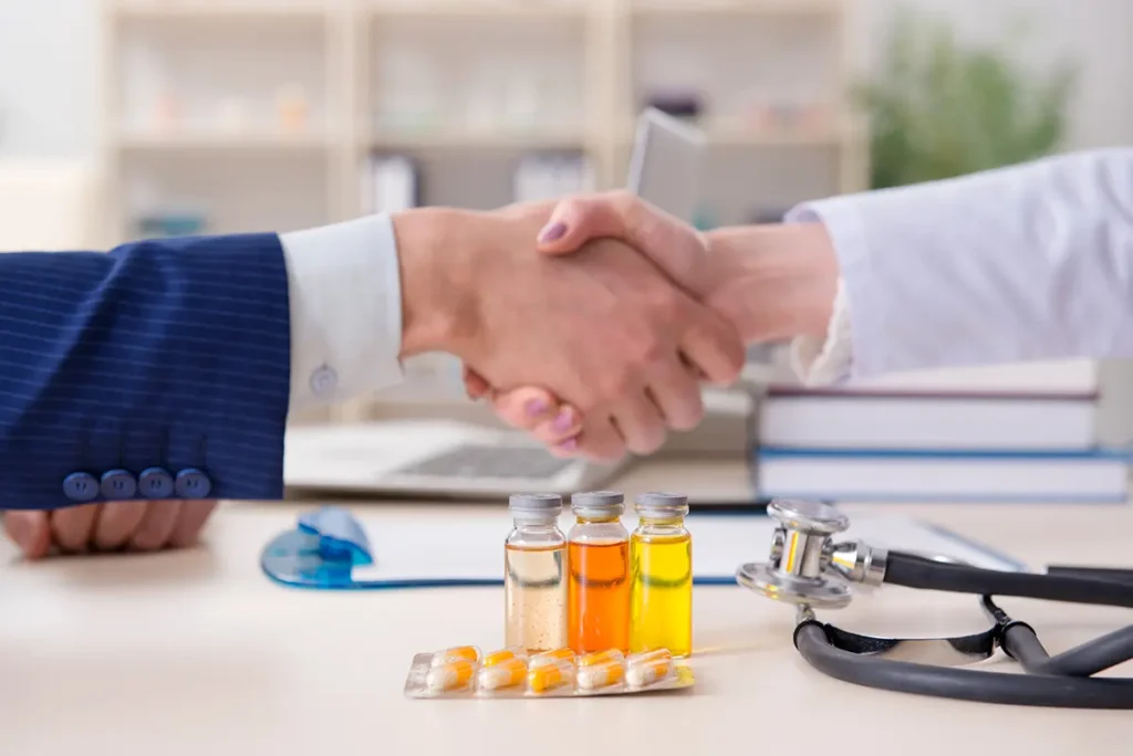 Two medical professionals shaking hands over a table with a stetehscope and severals vials of medicine as well as a foil-pack of tablets. Healthcare fraud concept.